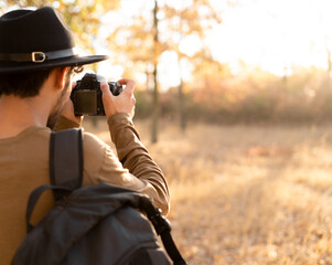 Male tourist photographing nature. Man in hat with slr camera and backpack hiking in autumn forest. - obrazy, fototapety, plakaty
