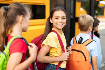Portrait Of Smiling Girl With Backpack Boarding Yellow School Bus With Classmates