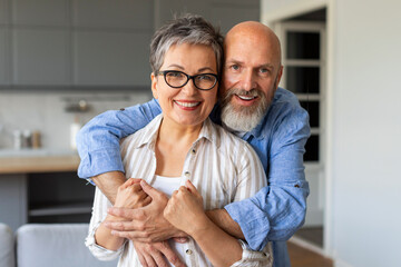 Portrait Of Loving Senior Spouses Embracing At Home And Smiling At Camera