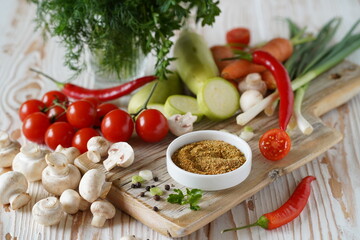 spices are ground in a small plate on a wooden board. vegetables around: zucchini, tomatoes, champignons, carrots, chili peppers, and greens in a glass, close-up, light background