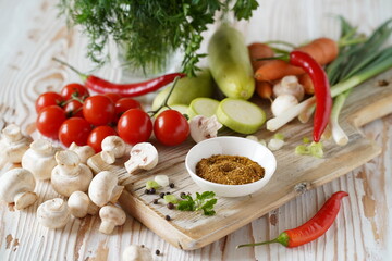 spices are ground in a small plate on a wooden board. vegetables around: zucchini, tomatoes, champignons, carrots, chili peppers, and greens in a glass, close-up, light background