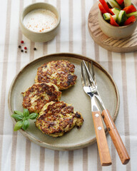 vegetable pancakes on a ceramic plate, cutlery with wooden handles, sauce pan with sauce next to it, and a bowl with vegetables, light background
