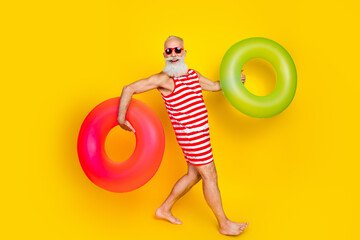 Full length photo of funny carefree senior man wear red striped swimsuit walking holding inflatable...