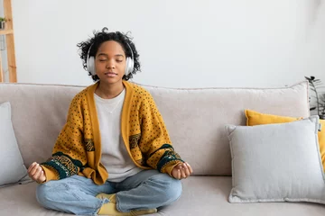 Rolgordijnen Yoga mindfulness meditation. Young healthy african girl practicing yoga at home. Woman sitting in lotus pose on couch meditating smiling relaxing indoor. Girl doing breathing practice. Yoga at home © Юлия Завалишина