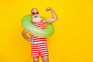 Photo of cool positive senior man wear red striped swimsuit showing muscles holding inflatable circle empty space isolated yellow color background