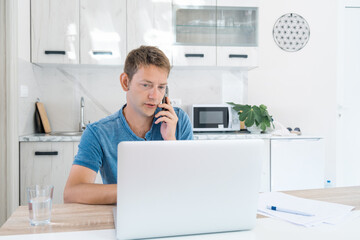 Serious young businessman trader talking on phone call while work on laptop at home. Male...