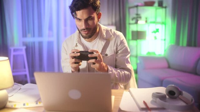 Happy excited young guy with joystick controller playing video games on laptop at late night Smiling young man have fun relax after hard work day or study indoors Game addiction concept