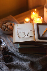 Bowl of cookies, cup of tea or coffee, plate of blueberries, dry oranges, wool yarn, pine cones, books, reading glasses, tablet, pen, soft blanket and lit candles on the table. Winter hygge at home.