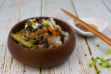 Vegan rice and vegetable stir fry in wooden bowl on white wood table. Chopsticks, restin on a small plate with sesame seeds