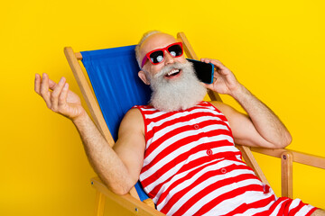 Portrait of funny cool pensioner man lying on sunbed at summer resort and phone calls abroad isolated over yellow color background