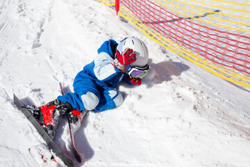 preschooler boy in a helmet, goggles, skis and winter overalls learns to ski, fell and lies on the...