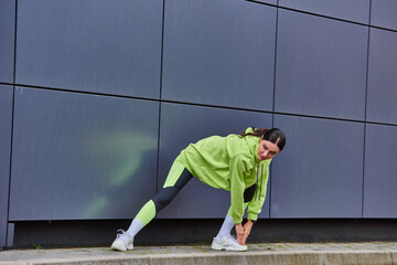 energetic woman in hoodie and leggings warming up near grey wall outdoors, motivation and sport