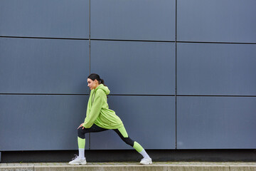 young sportswoman in hoodie and leggings stretching legs near grey wall outdoors, energy and sport - Powered by Adobe