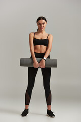 full length of young sportswoman in active wear holding fitness mat and standing on grey background