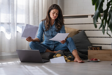 Stressed young woman holding financial papers sitting on the floor concerned about her finances - obrazy, fototapety, plakaty