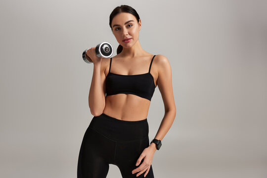 pretty and strong woman in active wear lifting dumbbell while working out on grey backdrop, banner