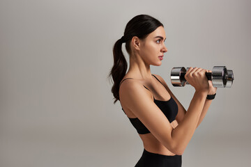 side view of sportive young woman in black active wear lifting dumbbells on grey background
