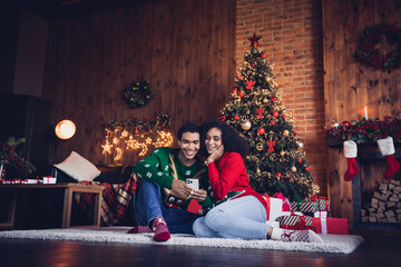 Photo of two people friends in comfort cozy flat room indoors with lights decoration browsing smart cell gadget for christmas events