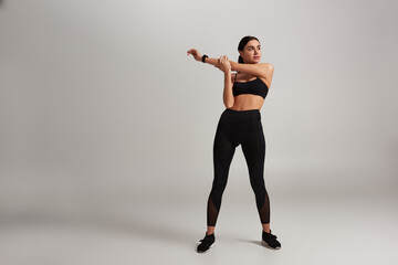 Fototapeta na wymiar young sporty woman in black sportswear with fitness tracker on wrist stretching on grey backdrop