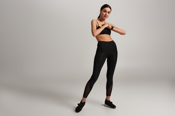 young sporty woman in black sportswear with fitness tracker on wrist exercising on grey backdrop