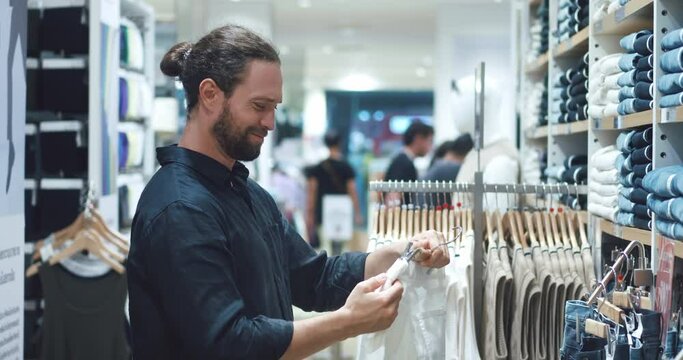 Young man checks price tags in clothing store. buyer checks price is surprised by the high checks price of a product. man sighs in bewilderment and hangs the goods back side view of bearded man