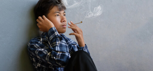 Asian boy sitting in front of dark wall of the toilet and smoking seriously, soft and selective focus, homeless and drug addiction concept. - obrazy, fototapety, plakaty