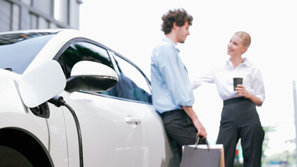 Progressive businessman and businesswoman leaning on electric car connected to charging station...