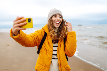 Happy woman in a bright jacket on the seashore with a phone in her hands. Young tourist woman takes...