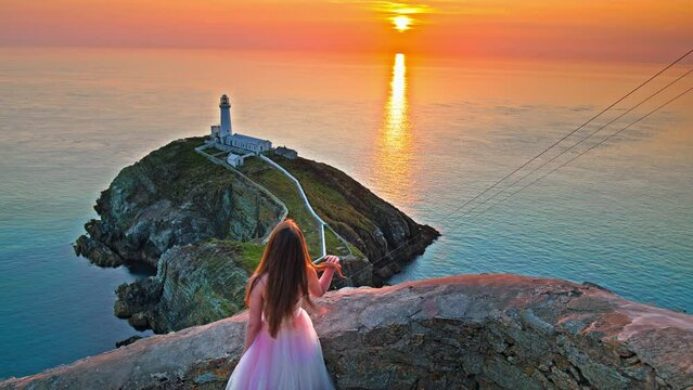 Beautiful woman in a dress visits South Stack Lighthouse at golden sunset in Holy Island, Wales. A tourist in a prom wedding dress walks at the Lighthouse with a footbridge over wild Atlantic ocean.