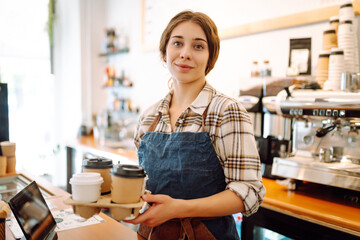 Female barista holding disposable takeaway cups. The young barista owner behind the bar gives out to-go orders. Business concept. Drinks to go.