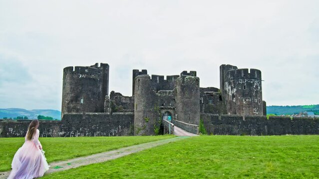 A beautiful woman in a princess dress visits Caerphilly Castle in Wales. A tourist girl in a long dress enjoys a moated fortress with an imposing gatehouse, a fortified dam and a tower.