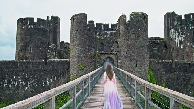 A beautiful woman in a princess dress visits Caerphilly Castle in Wales. A tourist girl in a long dress enjoys a moated fortress with an imposing gatehouse, a fortified dam and a tower.