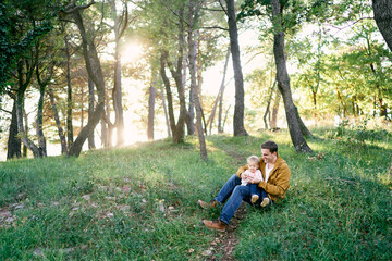 Naklejka na ściany i meble Little girl sits on her dad lap on green grass in a sunny forest