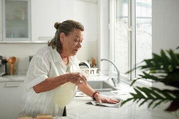 Aged woman wiping kitchen counter with cleaning detergent - obrazy, fototapety, plakaty