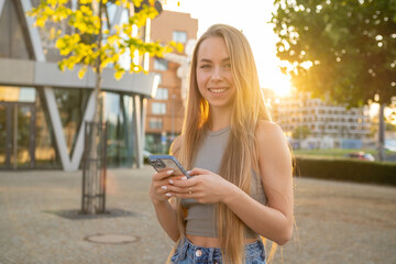 Joyful blonde lady with long loose hair brightly smiles looking in camera. Young woman happily laughs spending time in summer city park