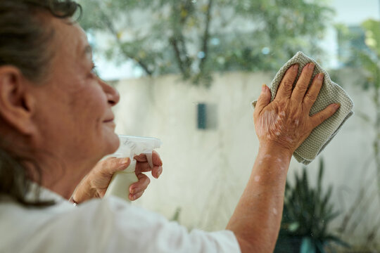 Cropped image of aged woman cleaning glass door with spray detergent