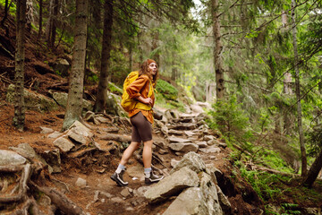Smiling woman traveler along a forest hiking trail in the mountains against the backdrop of nature. Young woman with backpack traveling outdoors. Hiking, active lifestyle.