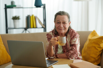 Cheerful elderly woman drinking tea and working on laptop at home