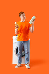 Young man with detergents near washing machine on orange background