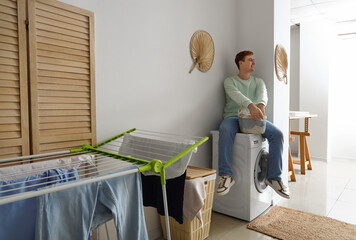 Young man with laundry basket sitting on washing machine at home - obrazy, fototapety, plakaty