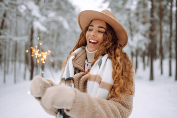 Fashionable woman in a stylish coat and hat holds sparklers in her hands in a snowy forest. A young female tourist in mittens with a New Year's spray bottle. The concept of holidays.