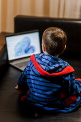 A little boy, a child in pajamas sits on a black leather sofa and looks at a laptop, playing games, watching interesting movies via the Internet. Modern childhood concept, copy space.