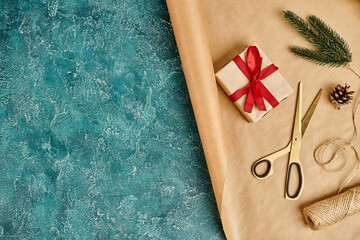 decorated gift box on craft paper and scissors near twine, pine cone and fir branch on blue backdrop - obrazy, fototapety, plakaty