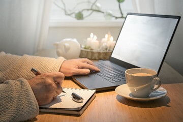 Hands of an older woman using laptop on a budget online shopping for Christmas presents and making notes on a table with advent candles, piggy bank and coffee, copy space - 674900213