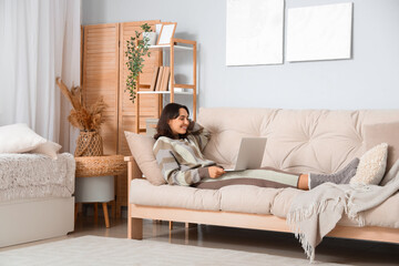 Young woman in warm sweater using laptop on sofa at home - obrazy, fototapety, plakaty