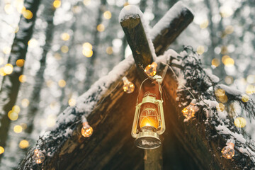 Primitive survival shelter in winter forest decorated with garlands and light of kerosene lamp....