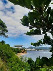 View of the Museum of Contemporary Art (MAC) in Niterói, Rio de Janeiro, Brazil, with Icaraí Beach with its tall buildings in the background, blue sky with clouds and foliage in the foregrou
