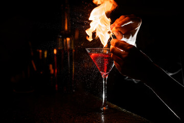 Transparent stemmed cocktail glass with brown alcoholic drink and flame above it stands on bar...