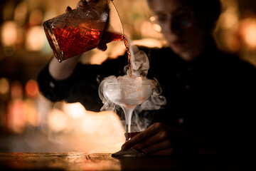 Female hands of a bartender begin to pour a cocktail from a mixing glass into a stemmed glass with...
