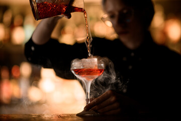 Female bartender enjoying making a smoky cocktail, pouring it into a stemmed glass with ice
